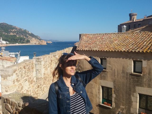 JaneRoa with tits covered, wearing a denim jacket and striped shirt, shielding eyes with hand, standing in front of a stone building with a tiled roof, by the sea.