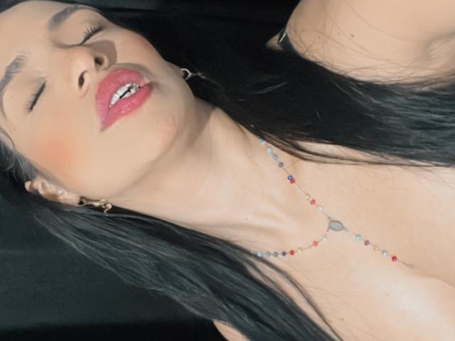 HeraDiosa with eyes closed, pink lipstick, black hair, gold hoop earrings, and a beaded necklace, her head tilted back, showing her bare shoulders and a hint of her tits.