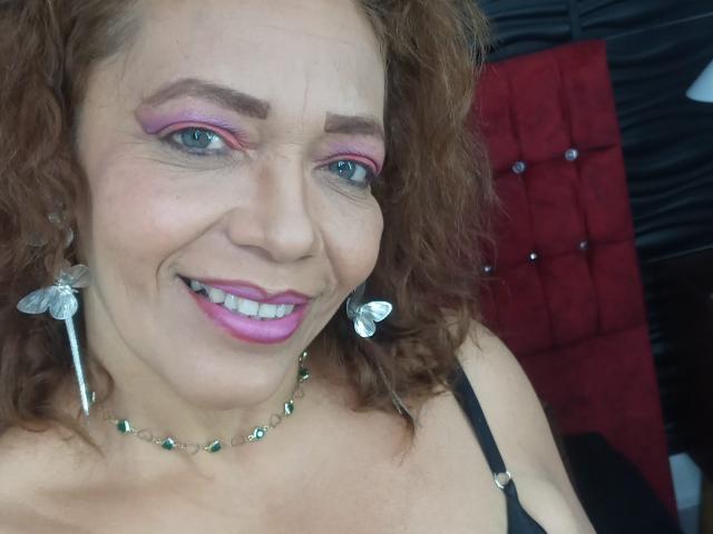 ZoeGreyX has curly brown hair, pink eyeshadow, green eyes, and pink lipstick. She's smiling with butterfly earrings, green necklace, and black strap showing her tits.