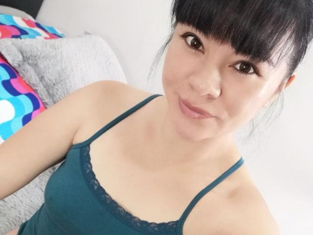 DayannaSquirt has black hair, brown eyes, and is wearing a teal lace-trimmed tank top, showing her small tits and a subtle smile in a selfie.