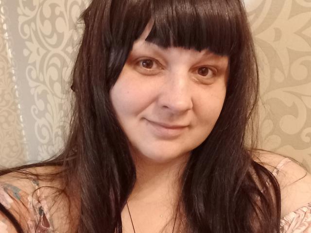 LuNora with straight black hair and bangs, smiling slightly, pale skin, brown eyes, wearing a pink floral top, showing a small tattoo on her left shoulder.
