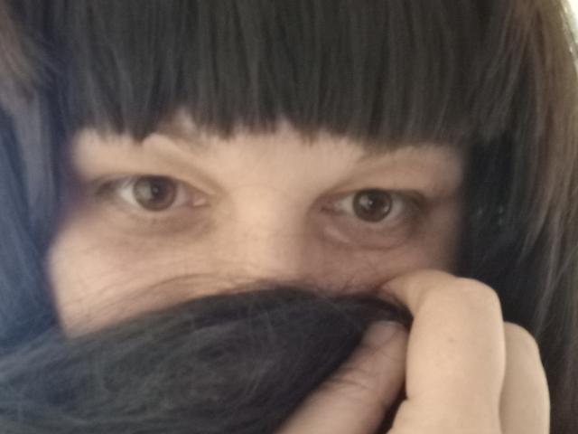 LuNora with dark, straight bangs, eyes looking forward, partially covering her face with black hair, using her right hand to hold her hair down. Her eyes are brown.