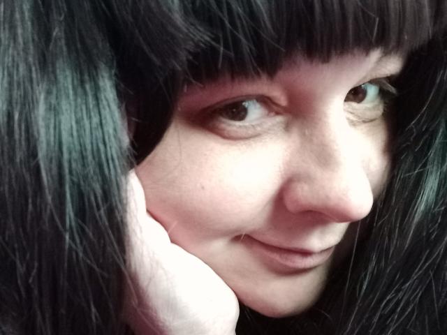 LuNora's face, with dark green-black hair, holding a pink phone to her ear, smiling slightly. Her brown eyes are focused, and she's showing a small nose and subtle lips.