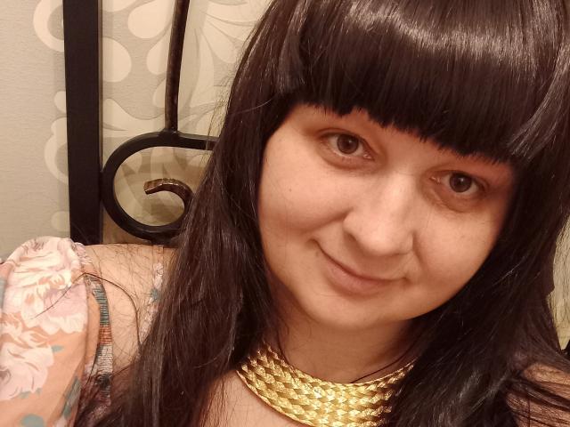 LuNora with black bangs, smiling, wearing a gold chain necklace, floral dress, and visible tattoo on her left arm, showing a cute face with brown eyes.