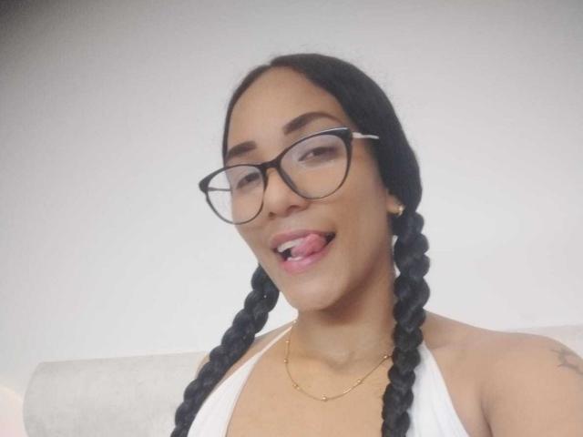 ThiainaRose a des tresses foncées, des lunettes et un collier en or. Elle a la langue tirée, montrant de petites dents blanches, et un tatouage sur son épaule droite. Elle porte un haut blanc..