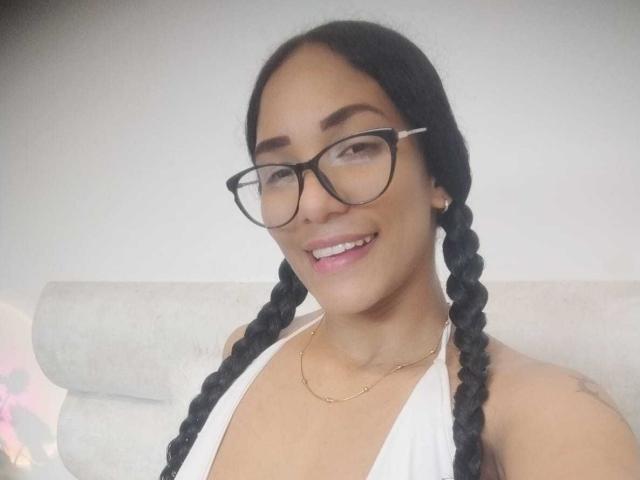 ThiainaRose con due trecce nere, occhiali, top bianco a collo alto, collana dorata, sorridente, che mostra i denti, pelle chiara, carina, attraente, seno piccolo, sfondo sfocato..