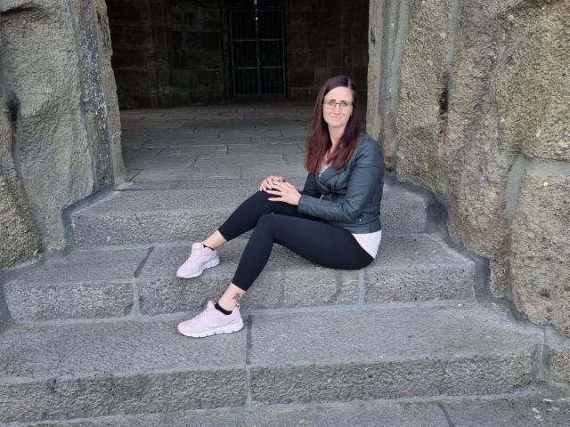NadjaNeah-hot, sitting on stone steps, wearing black leggings, pink sneakers, gray jacket, white shorts, with hands on knees, long brown hair, glasses, smiling, rock walls behind.