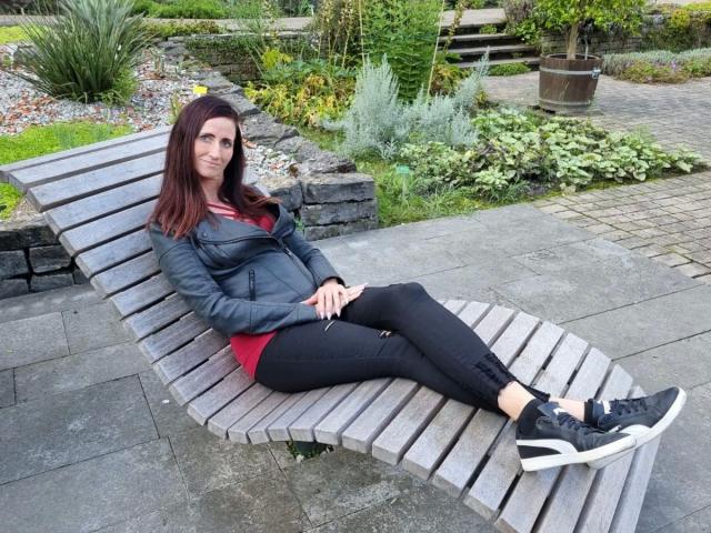 NadjaNeah-hot is on a wooden chaise lounge, wearing a black leather jacket, red shirt, black pants, and black sneakers, with her legs crossed, showing her ass.