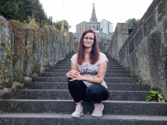 NadjaNeah-hot is squatting on stone stairs, wearing black pants, pink sneakers, and a white shirt, with tattoos on her arms, and a small smile, in a rocky, mossy setting.