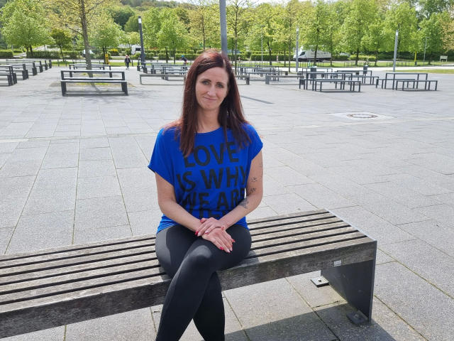 NadjaNeah-hot, with brown hair and blue "LOVE IS SWINGER" shirt, sits on a wooden bench, hands on knees, black leggings, small tits, tattoo on right forearm.