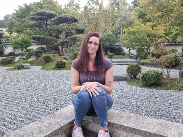 NadjaNeah-hot, sitting on a concrete ledge, has long red hair, glasses, a purple t-shirt, and blue jeans, with her hands on her knees, looking happy.