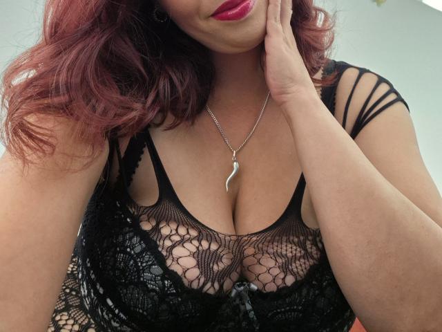 LadyHellena with red hair, wearing black lace lingerie that shows her big tits, touching her cheek with a hand, wearing a silver necklace with a curvy pendant.