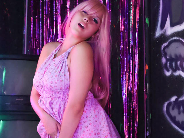Catalina Rogers, com cabelo rosa, vestindo um vestido floral rosa, inclinada para frente com a boca aberta, as mãos no bumbum, sob luzes neon roxas e rosas, com uma aparência sedutora..