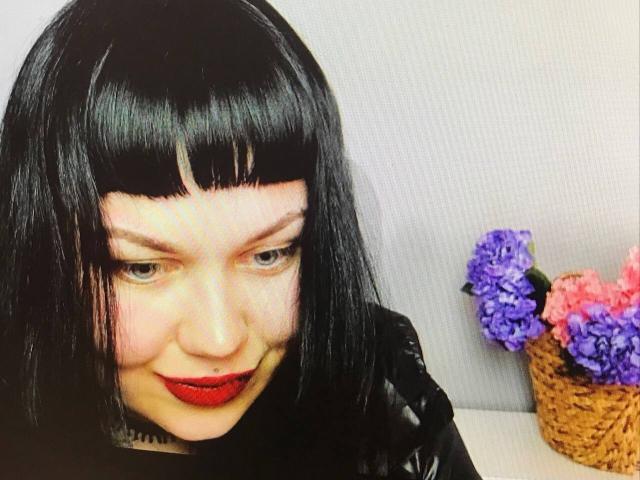 BetsiBlum mit schwartem Bob, rotem Lippenstift und heller Haut, die leicht grinst. Sie hat lila und rosa Blumen in einem gewobenen Korb auf der rechten Seite..