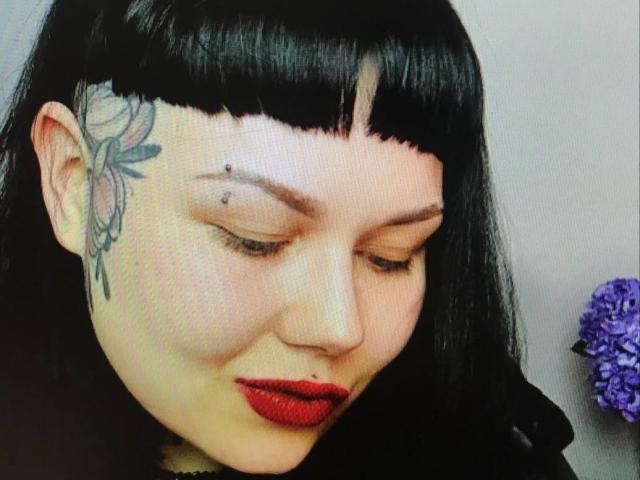 A BetsiBlum tem franjas pretas, batom vermelho e uma tatuagem de uma flor na têmpora direita, além de um piercing no rosto. Ela está olhando para baixo, com flores roxas ao fundo..