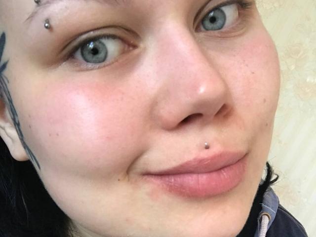 Il viso di BetsiBlum, con occhi azzurri, naso e labbro forati, mostra un leggero sorriso con labbra rosa, leggermente dischiuse, e un tatuaggio nero sul lato sinistro..