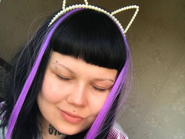 A BetsiBlum tem cabelo preto e roxo, com orelhas de gato, olhos fechados, um piercing no nariz e um piercing no lábio. Ela está sorrindo, com um pequeno piercing no nariz e vários piercings faciais..