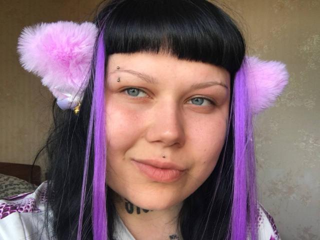 BetsiBlum ha i capelli neri e viola, con orecchie da gatto rosa e pelose, un anello al naso, un piercing al labbro e tatuaggi sul collo, con gli occhi azzurri e un'espressione accigliata..