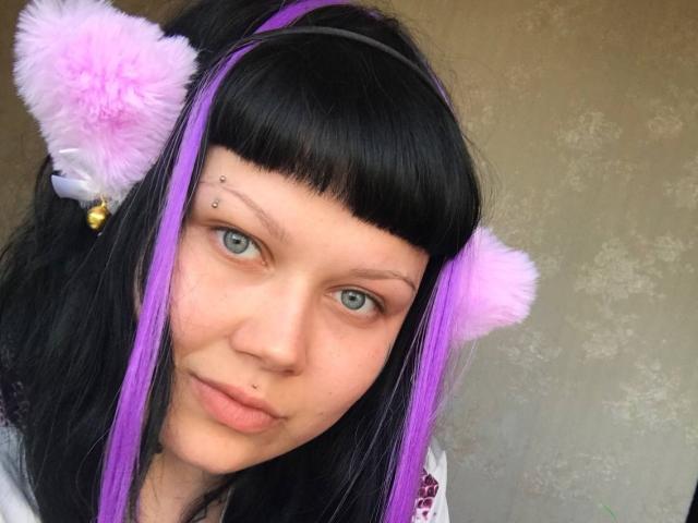 BetsiBlum, com cabelo preto, mechas roxas e orelhas de gato fofas cor-de-rosa, tem um piercing no nariz, um piercing no lábio e uma pinta no rosto, e está olhando com olhos azuis..