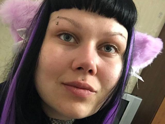 BetsiBlum, avec des cheveux noirs et violets, des oreilles de chat violettes, un piercing noir au nez et un petit piercing noir au sourcil, sourit à la caméra. Ses yeux sont bleus et elle a les lèvres roses..