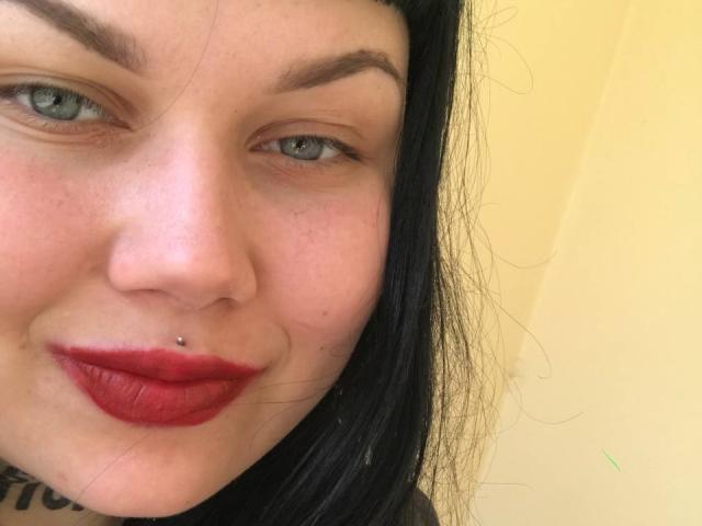 O rosto de BetsiBlum, com pele clara, olhos azuis, sobrancelhas pretas, batom vermelho, piercing no nariz preto, cabelo preto, um leve sorriso, sardas e uma tatuagem visível perto da parte inferior esquerda..