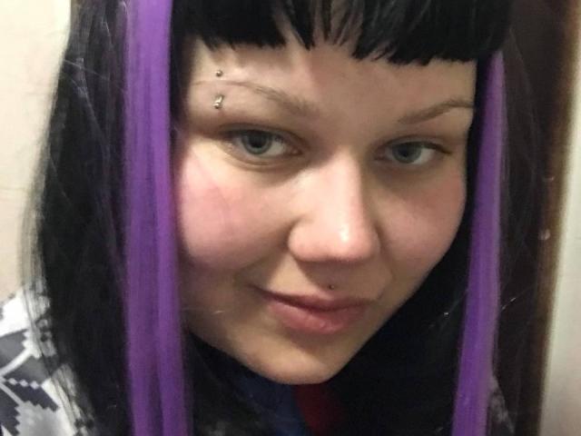 A BetsiBlum, com cabelo roxo, sobrancelhas pretas, piercing no nariz e no lábio, tem uma pequena perfuração na sobrancelha direita e um leve sorriso, mostrando uma pequena marca vermelha na bochecha..