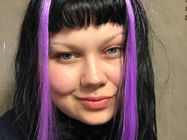 BetsiBlum mit schwarzen und lila Haaren, Pony, blauen Augen, Nasenring und Lippenpiercing, lächelt leicht. Sie hat einen süßen, punkigen Look mit ihren Haaren und Gesichtspiercings..