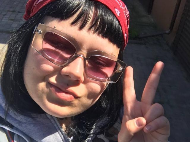 BetsiBlum, con i capelli neri, una bandana rossa e occhiali con una leggera sfumatura rosa, fa un segno di pace, ha un piercing al naso e un leggero sorriso, ed indossa una felpa grigia..