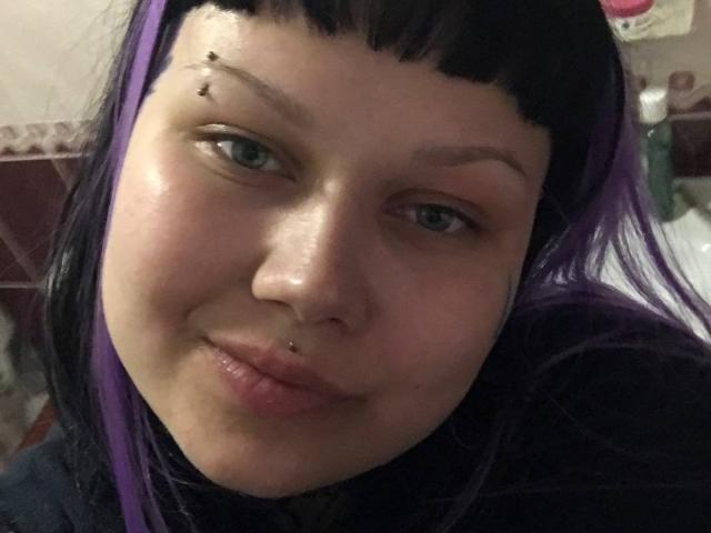 BetsiBlum, con i capelli neri e viola, piercing sul naso e sul labbro superiore, e un piccolo punto sulla fronte, sorride alla fotocamera. La sua pelle è pallida e luminosa..