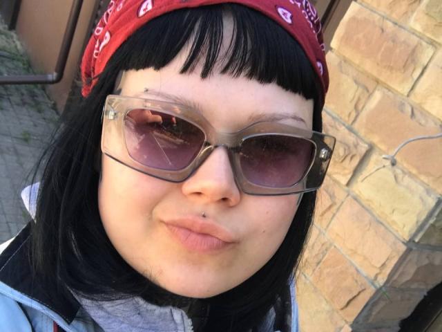 BetsiBlum, com cabelo preto, um lenço vermelho na cabeça, óculos de sol claros e um piercing no nariz, com os lábios curvados para baixo, vestindo uma jaqueta cinza com zíper, em frente a uma parede de tijolos..