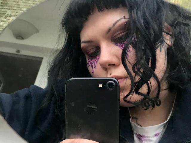 BetsiBlum, avec ses cheveux noirs et ondulés, son maquillage pour les yeux violet, et ses piercings au nez, tenant un iPhone noir, portant un sweat à capuche noir et un t-shirt blanc avec un motif violet..