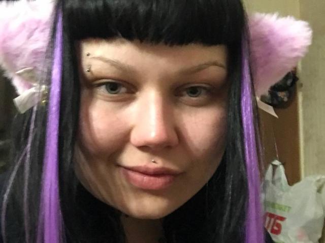 BetsiBlum a les cheveux noirs et violets, avec des oreilles de chat roses et duveteuses, un piercing au nez et une petite tache de naissance sur le visage. Elle sourit, avec une légère ride au menton..