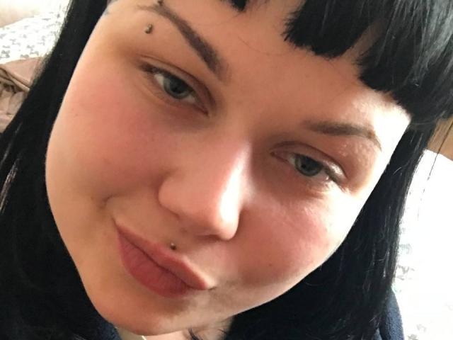 BetsiBlums Gesicht mit schwarzem, glattem Haar, blauen Augen und einem kleinen, schwarzen Schönheitsfleck oberhalb ihrer rechten Augenbraue. Sie trägt einen Nasenring und ein Lippenpiercing..