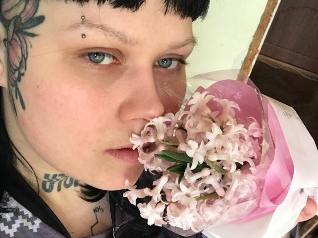 BetsiBlum, avec ses cheveux noirs, ses yeux bleus et ses tatouages, tient des fleurs lilas roses près de son nez. Elle a un piercing au nez et un tatouage floral sur sa joue gauche..