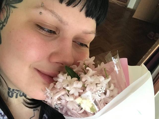 BetsiBlum, con cabello negro, perforación en la nariz y tatuaje, sosteniendo un ramo de lilas rosadas, sonriendo suavemente, mirando ligeramente hacia un lado, en una habitación con poca luz..