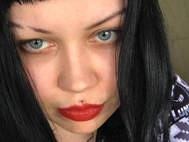 El rostro de BetsiBlum, con piel pálida, ojos azules, cabello negro y lacio, lápiz labial rojo y un piercing en la nariz. Su expresión es ligeramente seria, mirando directamente a la cámara..