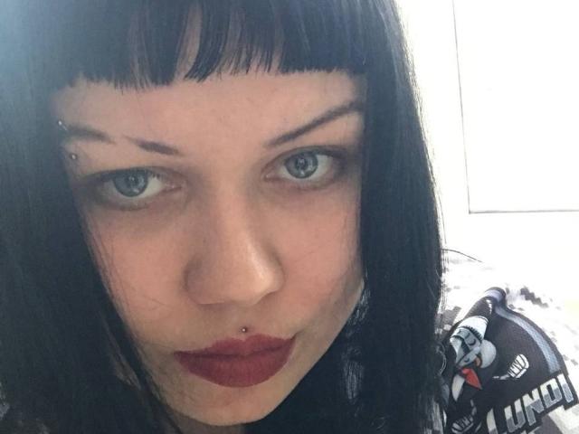 BetsiBlum con capelli neri e lisci, occhi azzurri, rossetto rosso, piercing al naso, piercing al labbro inferiore e sopracciglia alzate in un selfie. Le sue natiche non sono visibili..