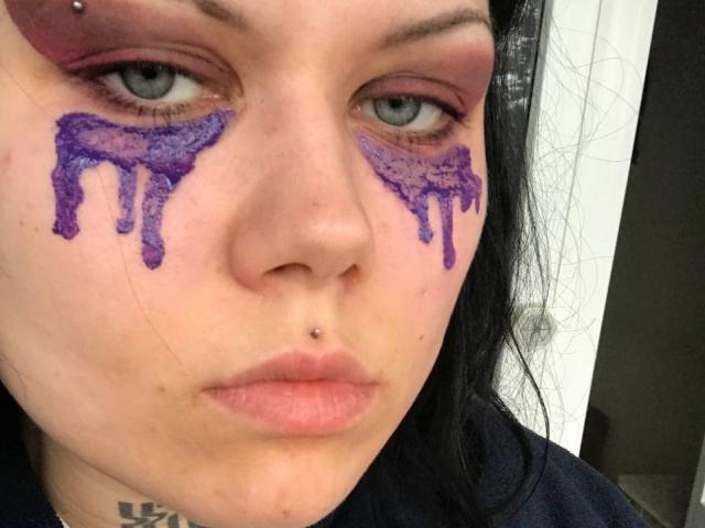 BetsiBlum hat lila Glitzer auf ihren Wangen, trägt rosafarbenen Lidschatten, hat ein Nasenpiercing und schwarzes Haar, sieht ernst aus mit einem leichten Schmollmund, hat blaue Augen und blasse Haut..