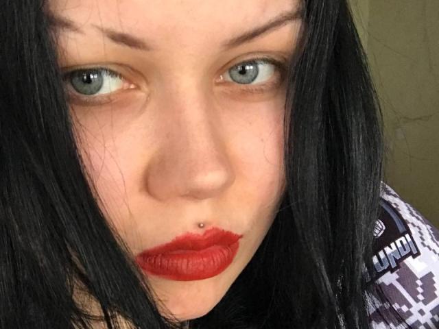 BetsiBlum tiene la piel pálida, ojos azules, cabello negro y lacio, lápiz labial rojo y un piercing en la nariz. Está mirando a la cámara con una expresión ligeramente fruncida..