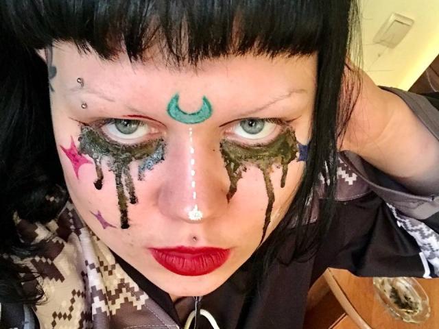 BetsiBlum's face is painted with green and black makeup, including dripping black lines and a green crescent on the forehead. She has red lipstick, black bangs, and a nose piercing.