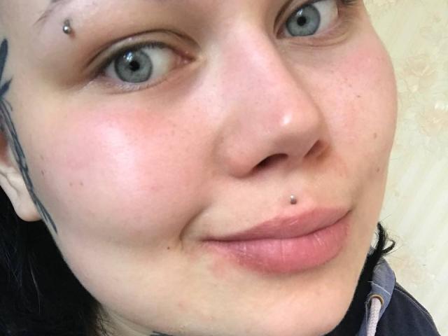 Het gezicht van BetsiBlum, met bleke huid, blauwe ogen, een zwarte wenkbrauw tattoo, een neusring, een lipring en licht open roze lippen. Haar zwarte haar is gedeeltelijk zichtbaar..