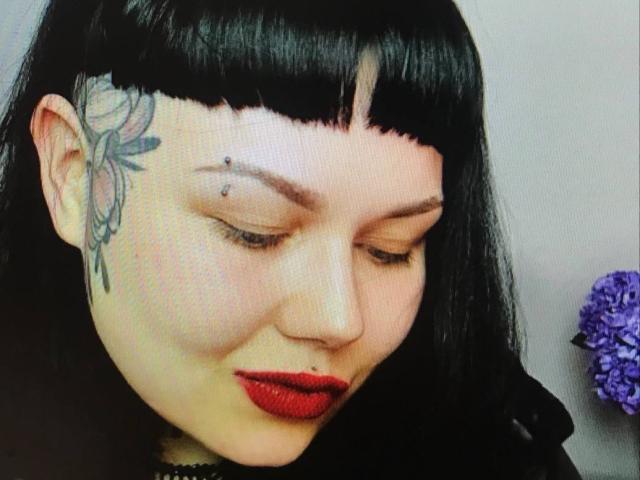 BetsiBlum heeft zwart haar met een pony, rode lippenstift en een bloementatoeage op haar linker slaap. Ze kijkt naar beneden met een lichte glimlach en laat een kleine neusring zien..