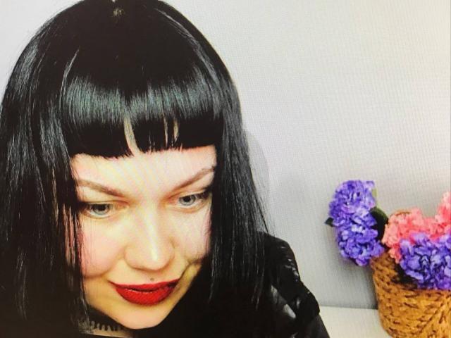 BetsiBlum, com um corte de cabelo bob preto, batom vermelho e um piercing no nariz, olhando ligeiramente para baixo, com uma cesta de vime cheia de flores roxas e rosas à direita..