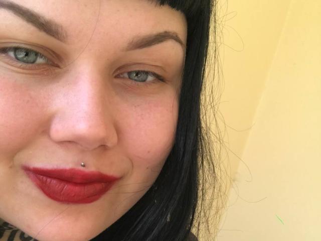 BetsiBlum has pale skin, black hair, green eyes, red lipstick, and a nose piercing. Her eyebrows are thick and she's smiling slightly.