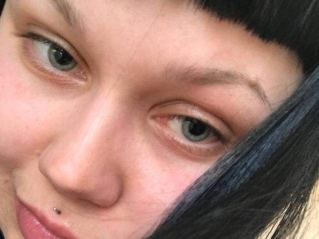 Het gezicht van BetsiBlum, met een neusring, zwart steil haar, groene ogen en een lichte glimlach, met een kleine puist boven haar rechter wenkbrauw..
