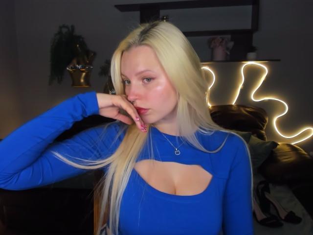 Alice Whitey, avec de longs cheveux blonds, un haut bleu découpé laissant entrevoir ses petites seins, une main près de son visage, des ongles roses et un collier, debout dans une pièce faiblement éclairée..