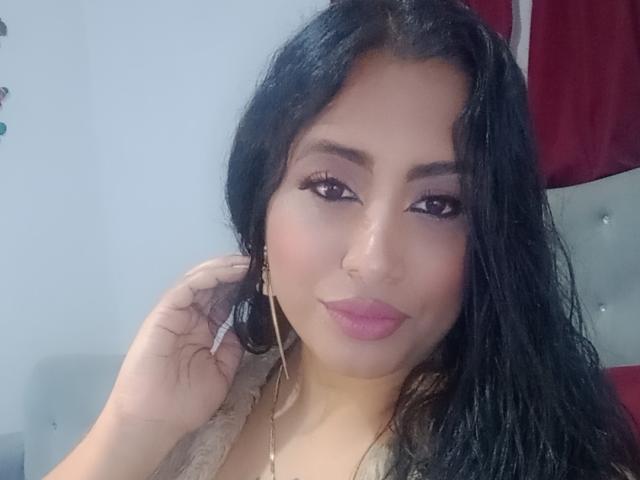 MariaCatalina with long black hair, brown eyes, pink lipstick, wearing a beige top, and large hoop earrings, smiling softly, touching her hair with her right hand.