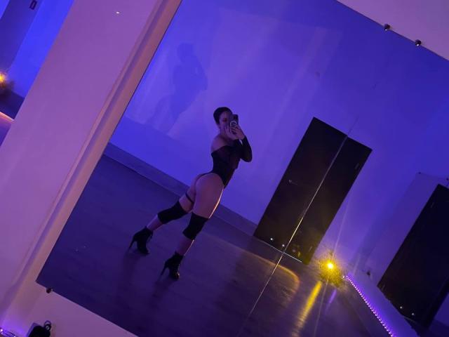 PaigePrado with long brown hair, winking, taking a selfie in a mirror, wearing a black bra, holding a phone with a ring, in a dimly lit room.