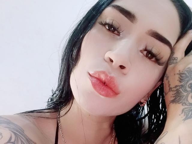 EvaMueller with long black hair, tattoos on arms and hands, wearing black fishnet lingerie, pouty lips, hand on chest, looking down at camera. Boobs partially visible.