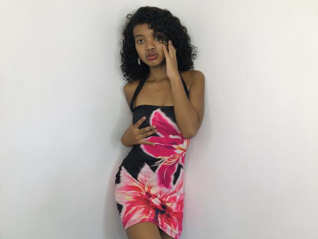 CandyMagicX with curly black hair, wearing a black halter dress with pink floral patterns, hands covering her tits, standing against a white wall, looking at the camera.